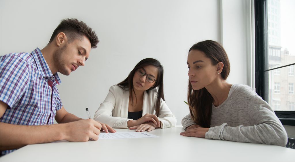 mesa com um homem de um lado, uma mulher de outro lado e uma mediadora entre os dois. O homem está assinando um papel.